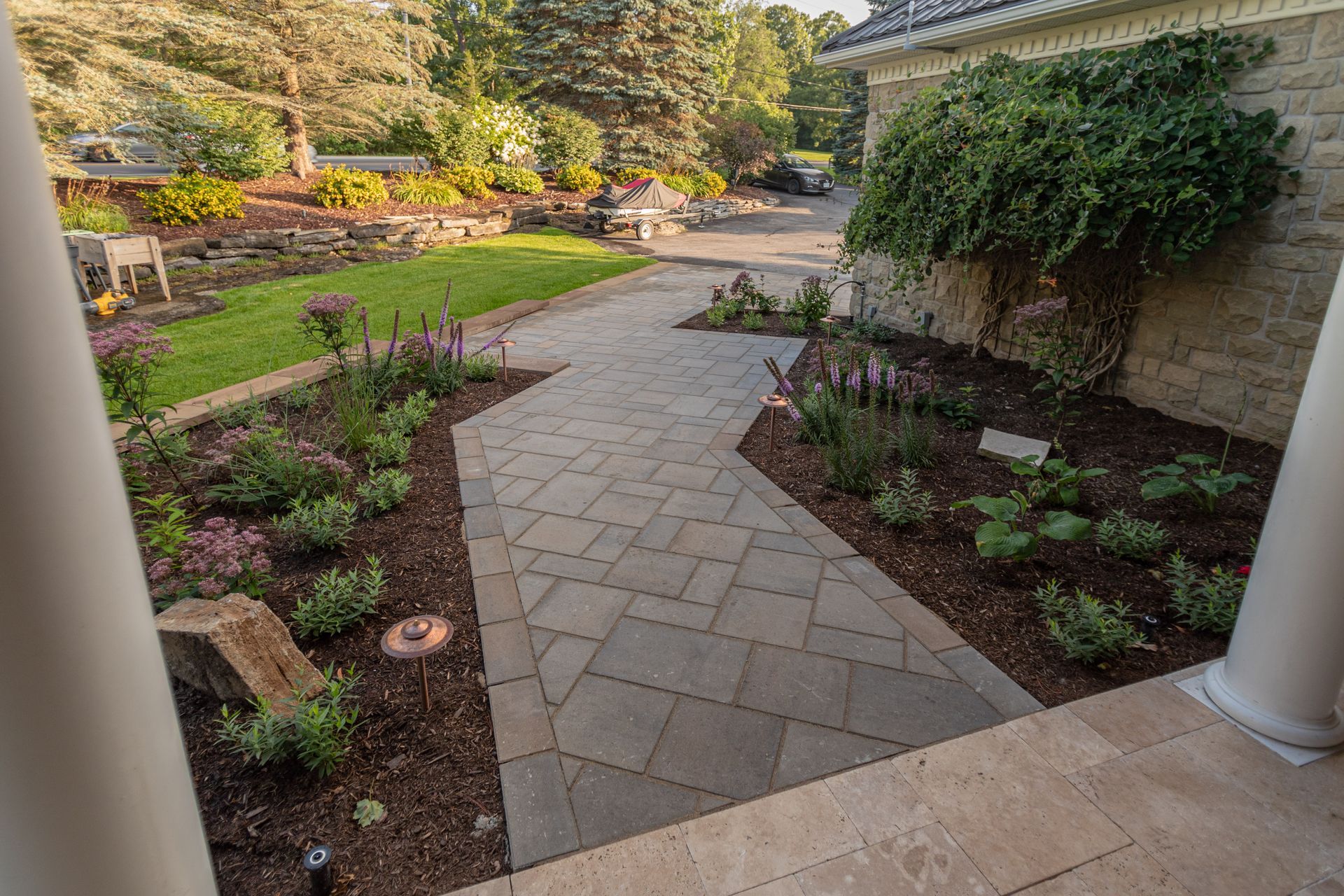 Brick pathway through landscaped garden, leading to lush greenery near a stone building.