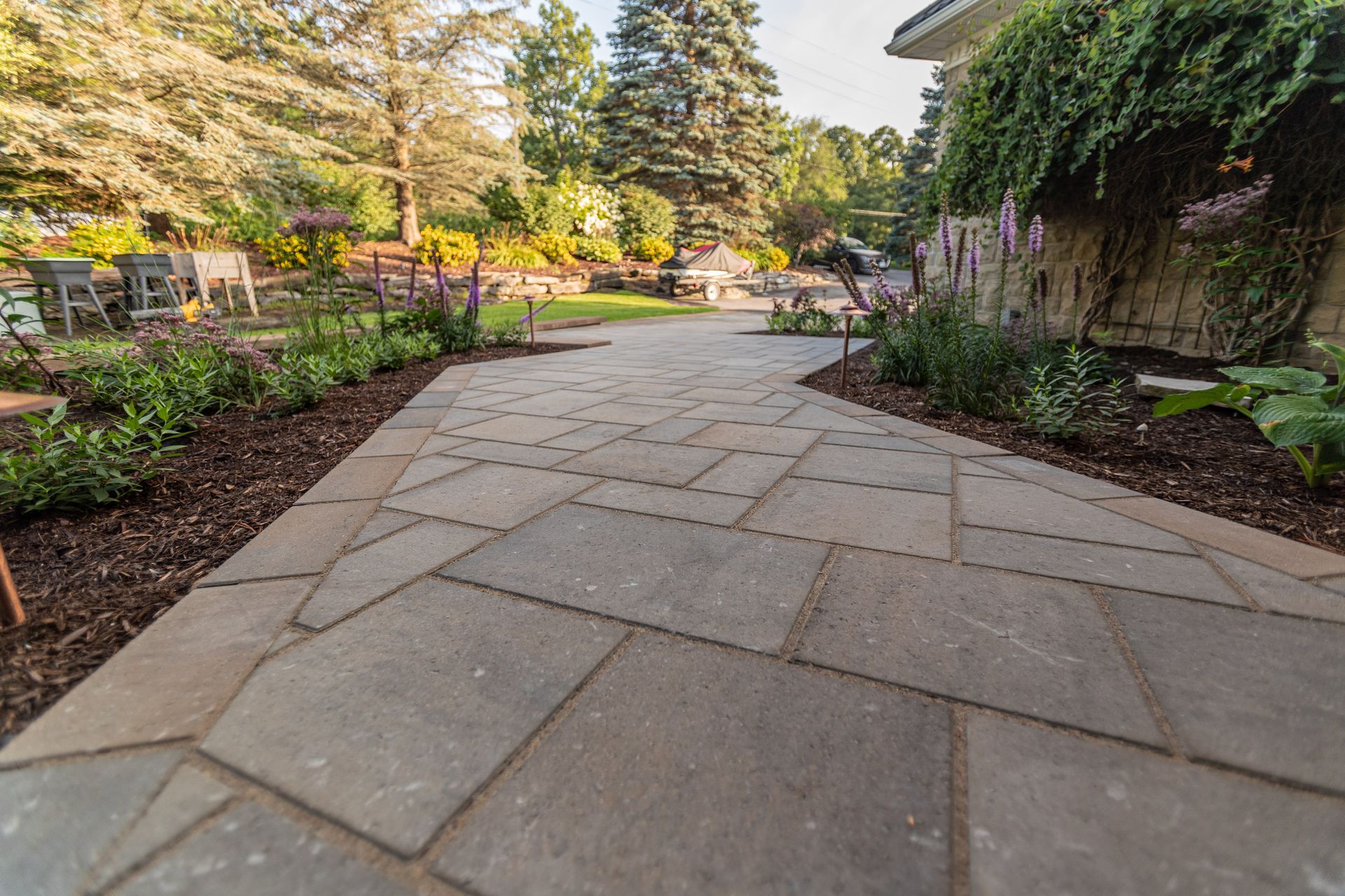 Stone pathway through a garden with landscaping. Brown mulch borders the path; trees and plants fill the background.