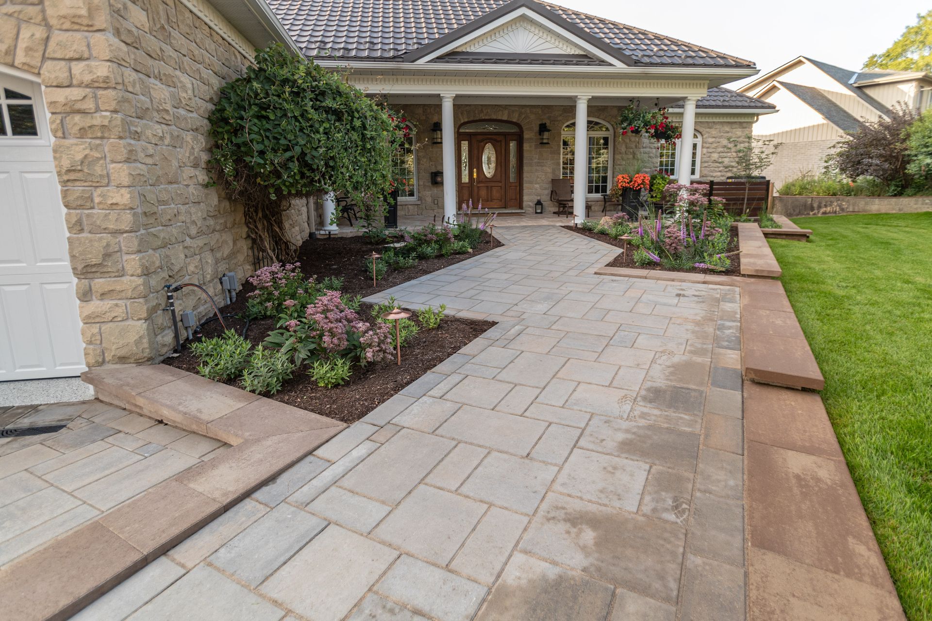 Stone walkway leading to a house with a covered porch, surrounded by flower beds and lawn.
