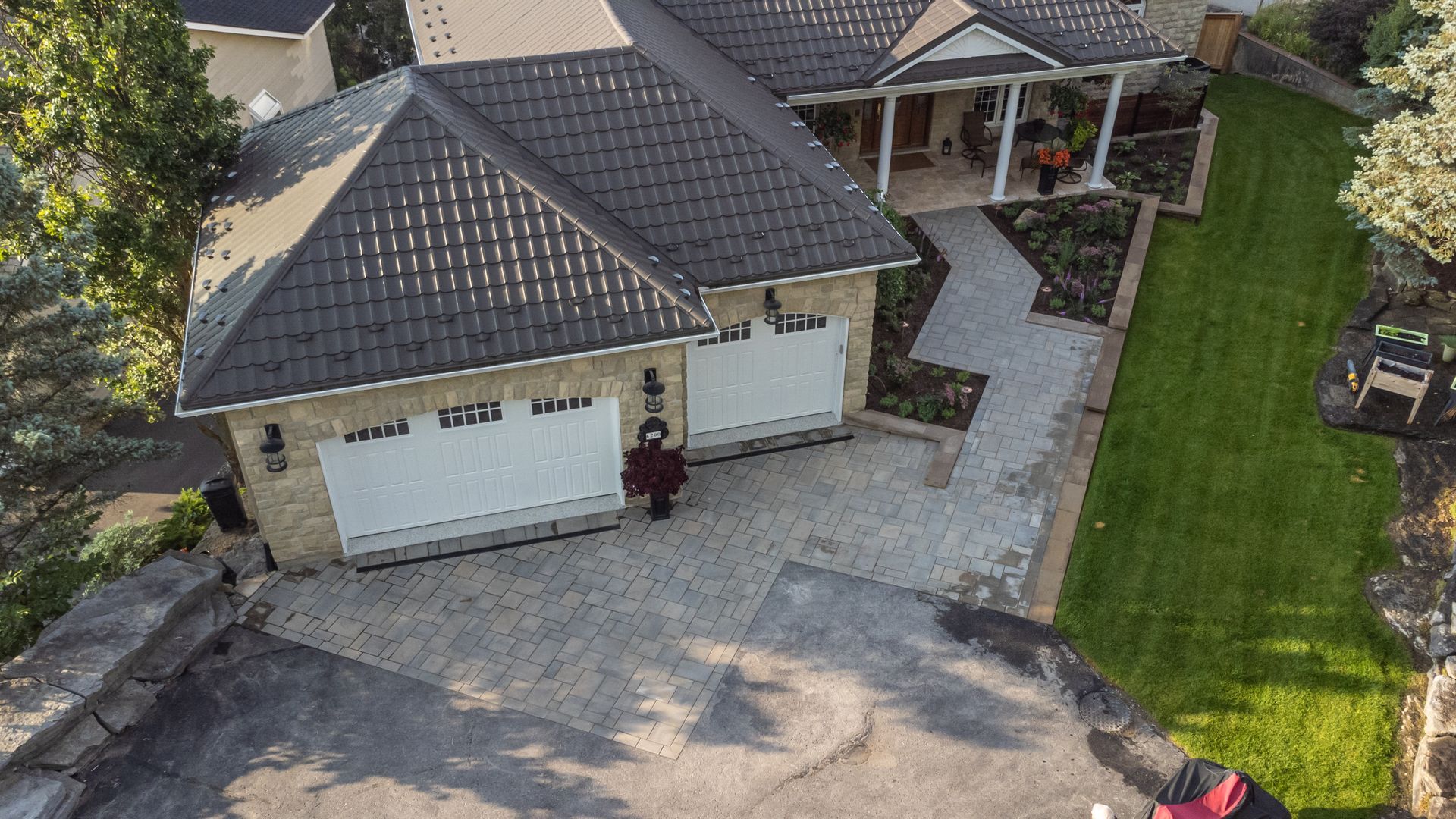 Two-car garage with light-colored doors, paving stone driveway, and a house with a porch and landscaped yard.