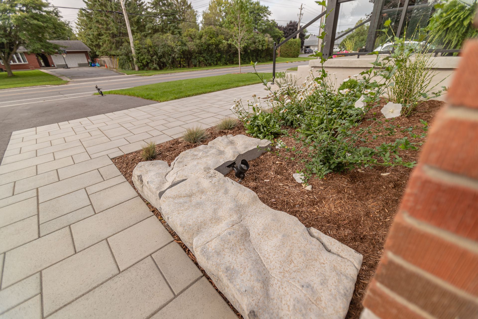 Paver walkway with a rock border, mulch, and greenery next to a brick wall. Street in background.