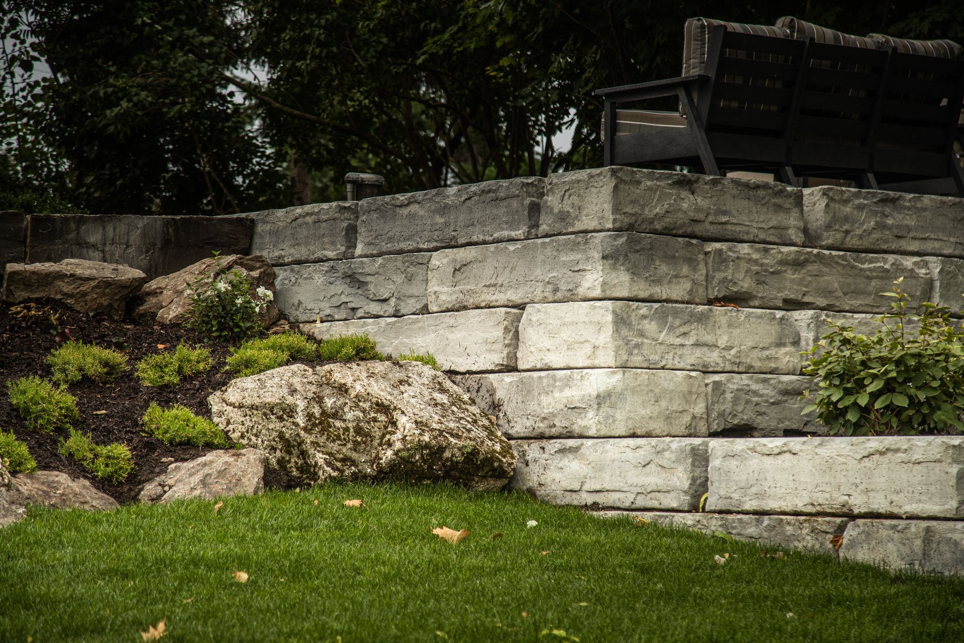 Stone retaining wall with green grass, landscaping, and a bench.