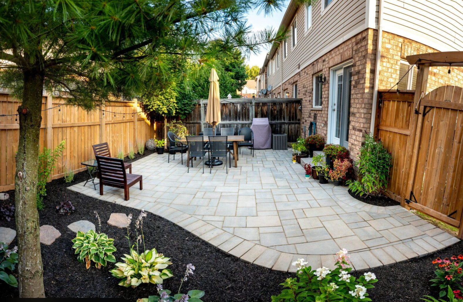 Interlock patio in a small backyard with curved edge, seating area, and surrounding garden beds.