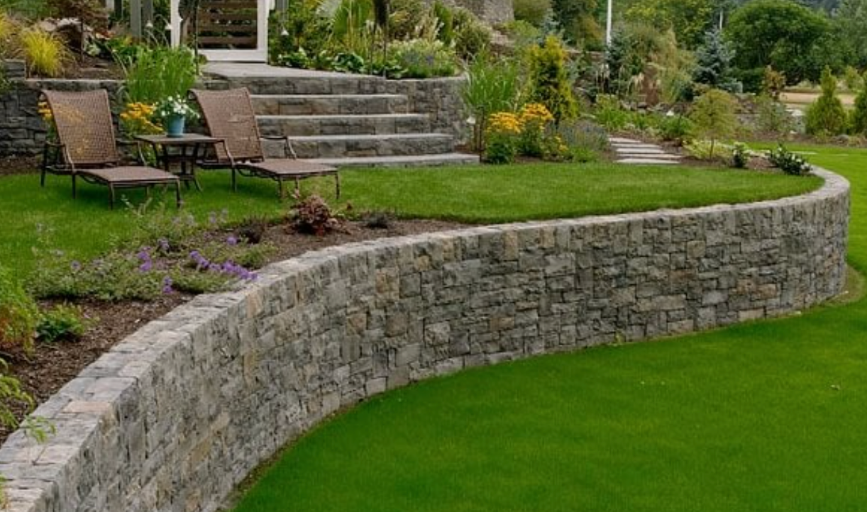 Curved natural stone retaining wall supporting a terraced lawn and patio steps with landscaped beds.