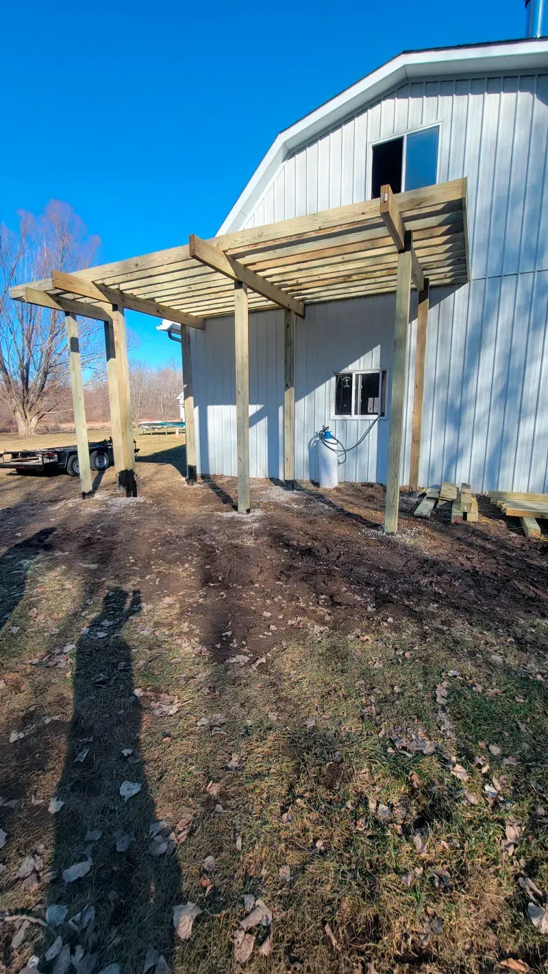 A wooden pergola is being built in front of a white barn.