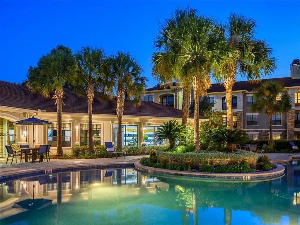 Luxury swimming pool and lounge area at dusk with palm trees and apartment buildings in the background.