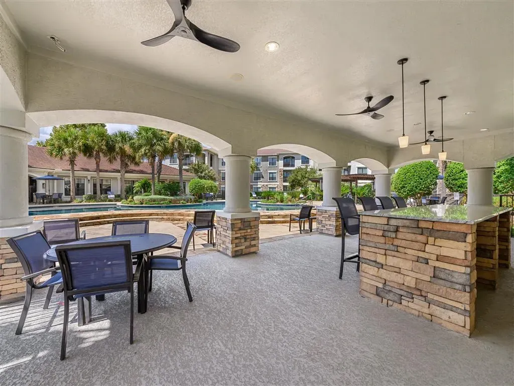 Outdoor covered patio with seating and a stone bar overlooking a pool and palm trees.
