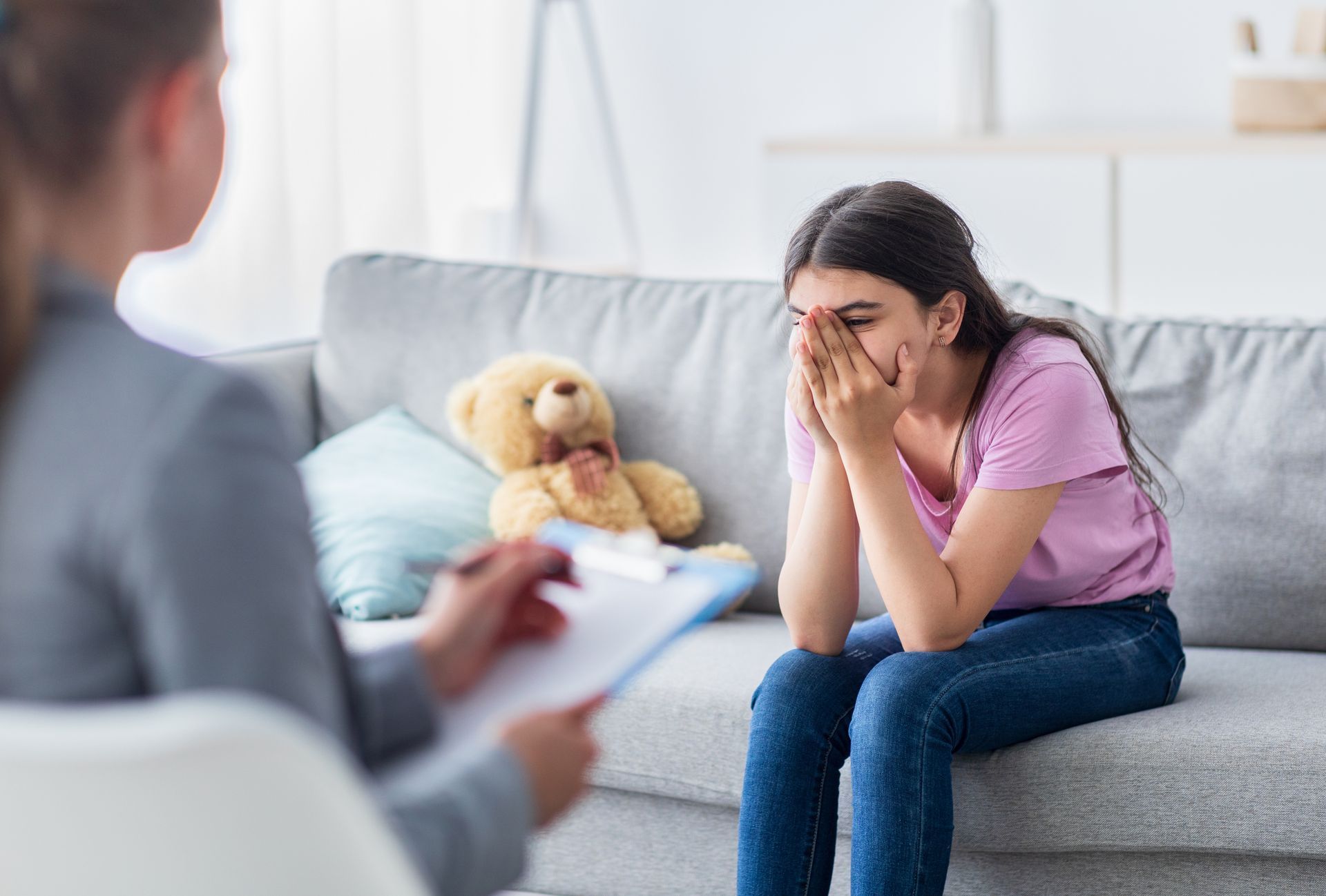 Woman sits on a couch with her face in her hands while a counselor listens nearby; teddy bear on the sofa.