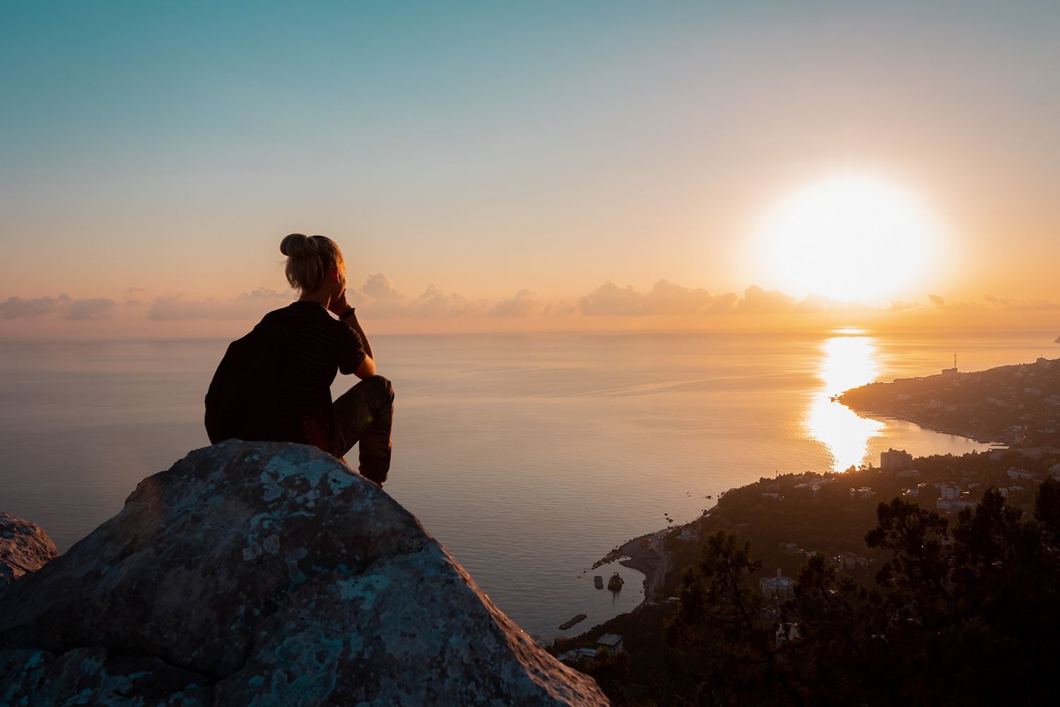 Woman seated on a rock overlooking ocean, watching sunset. Sun casts golden light; silhouette.