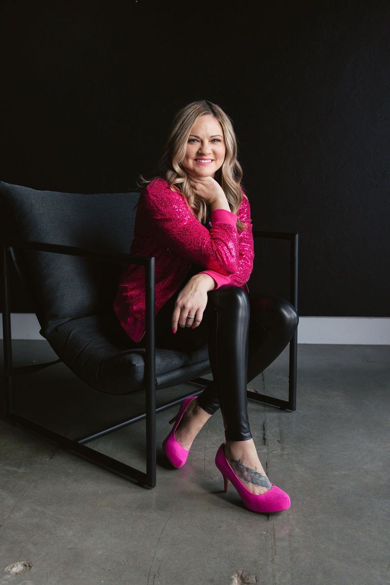 Woman in pink sequin jacket, leather leggings, and heels, sitting in a modern chair. Black background.
