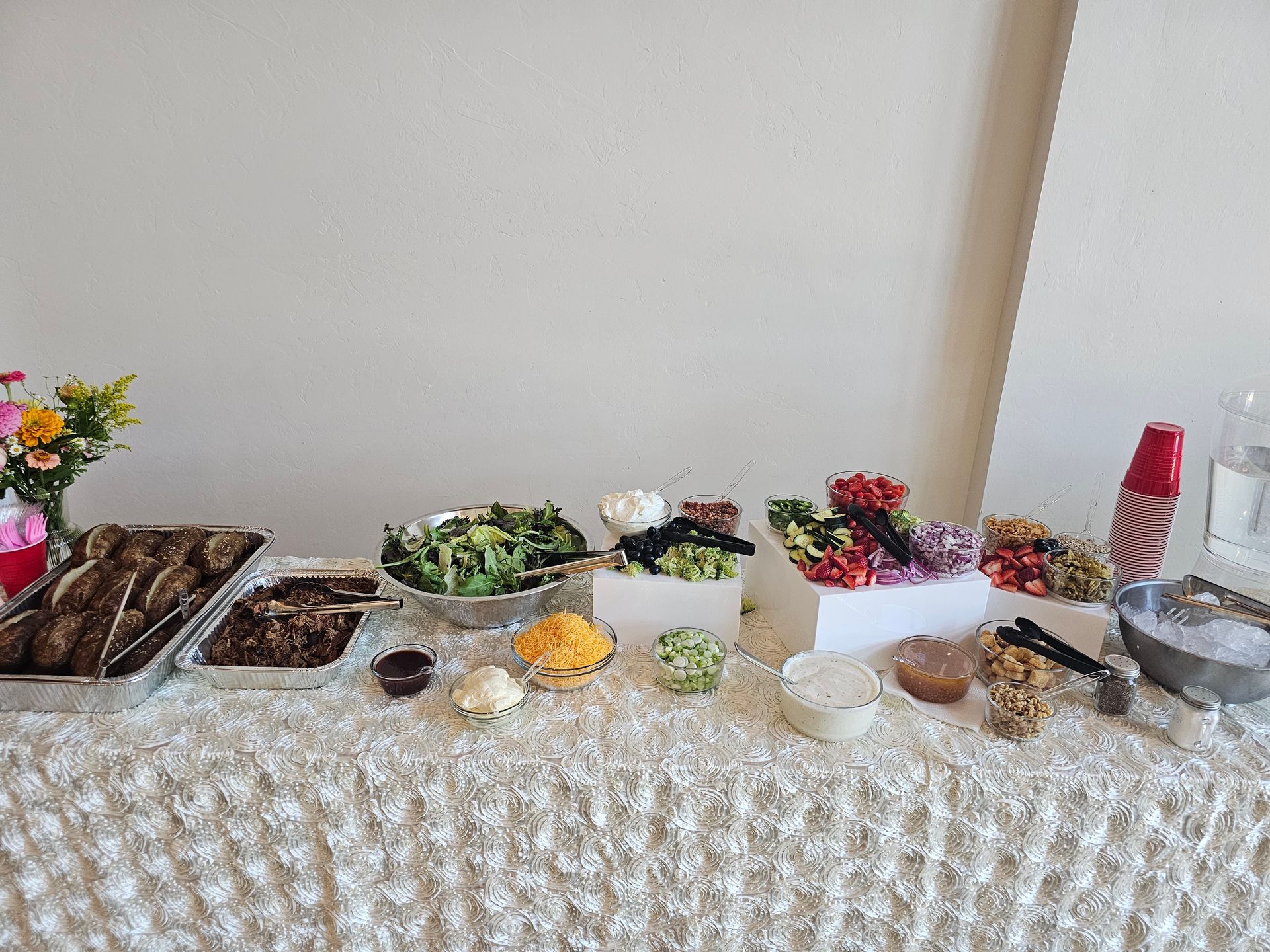 Buffet table with food items: meat, salad, toppings. Flowers and water pitcher in the background.