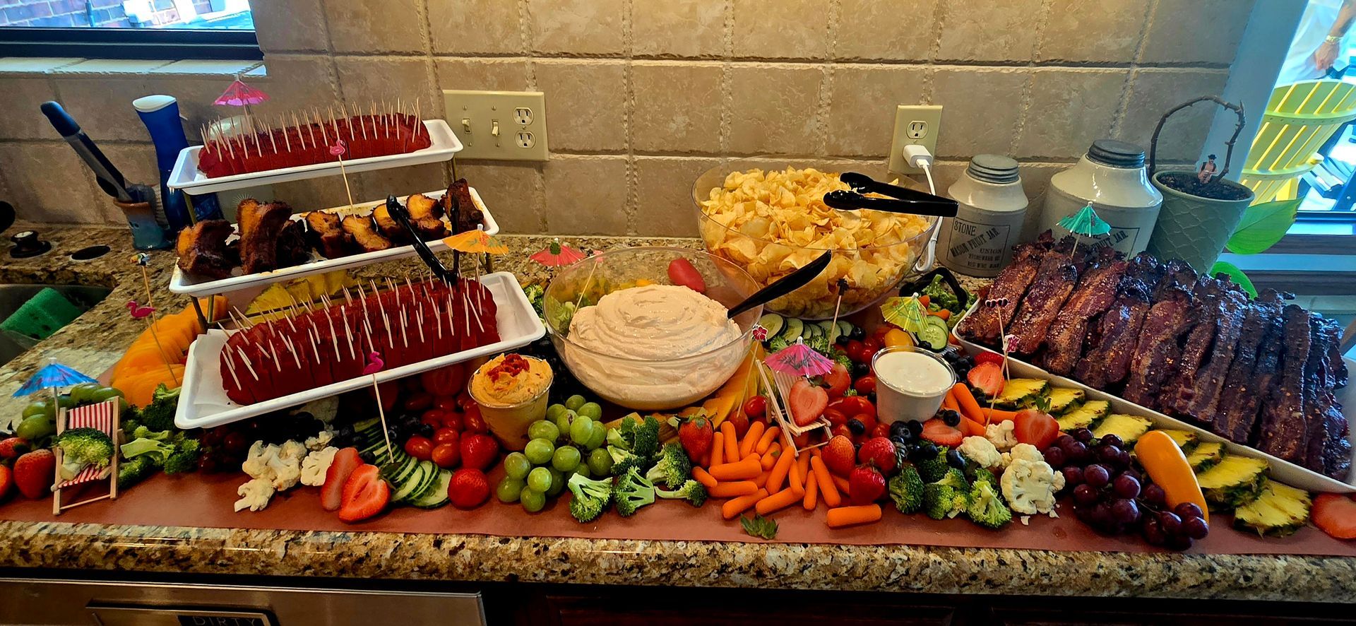 A lavish party spread with meat, veggies, dips, and desserts on a long serving board in a kitchen.