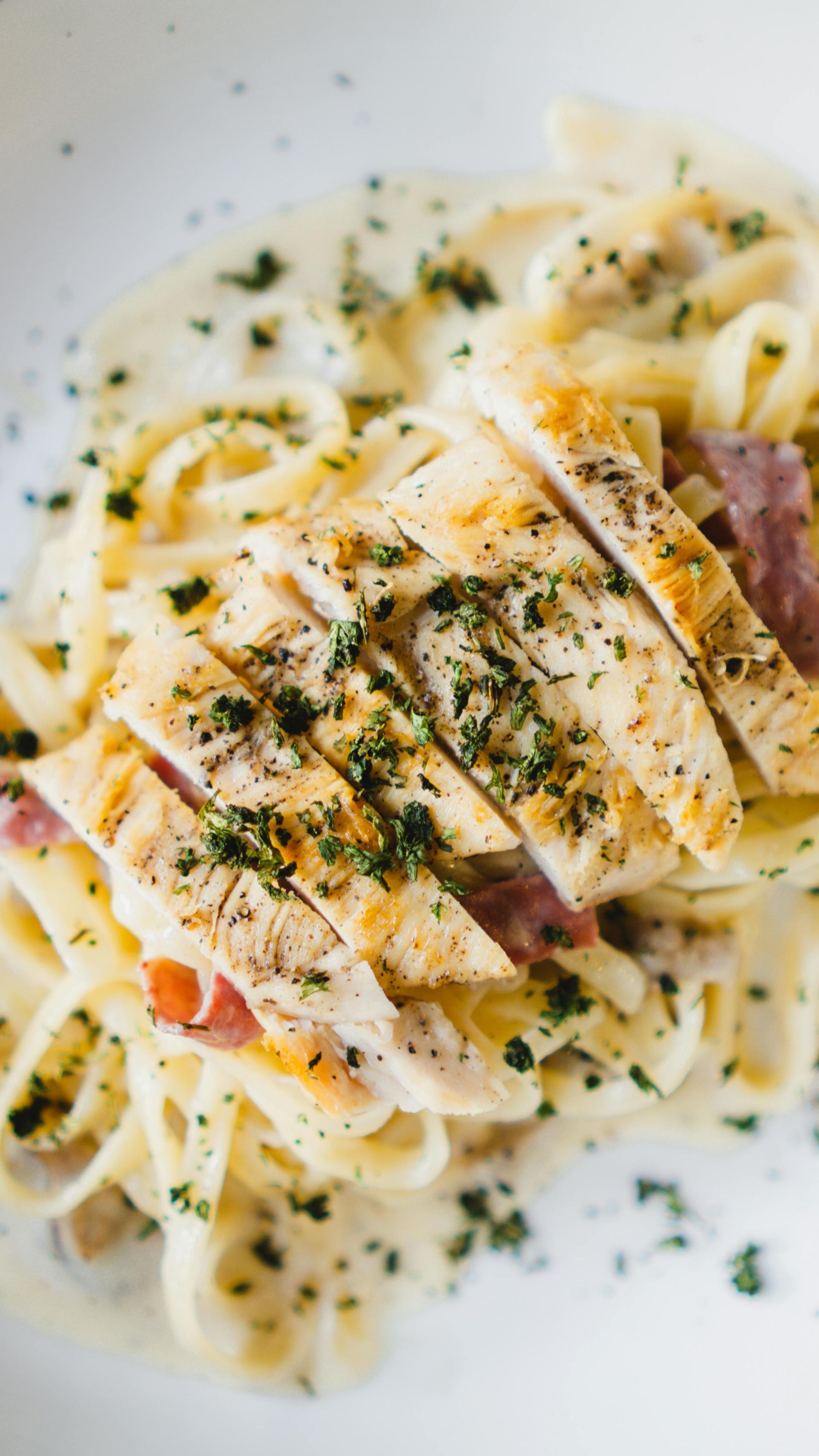 Fettuccine Alfredo with grilled chicken, topped with parsley, in a white bowl.