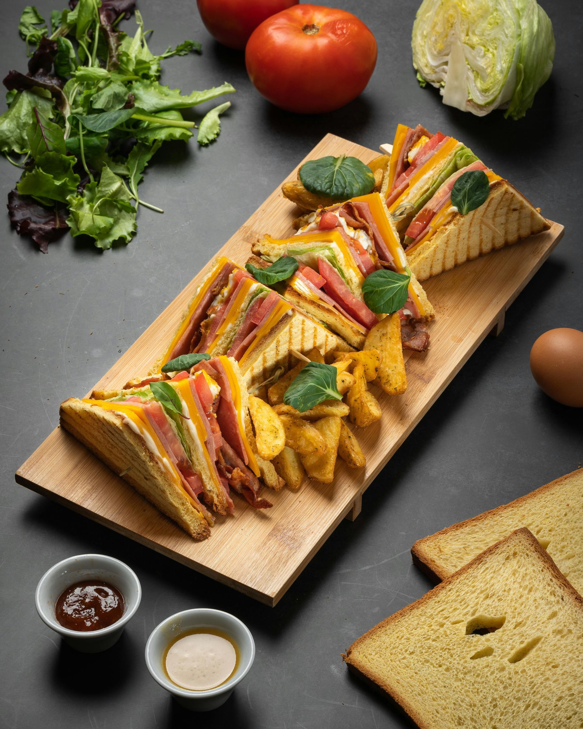 Club sandwiches on a wooden board, with tomatoes, lettuce, sauces, and bread slices on a dark surface.