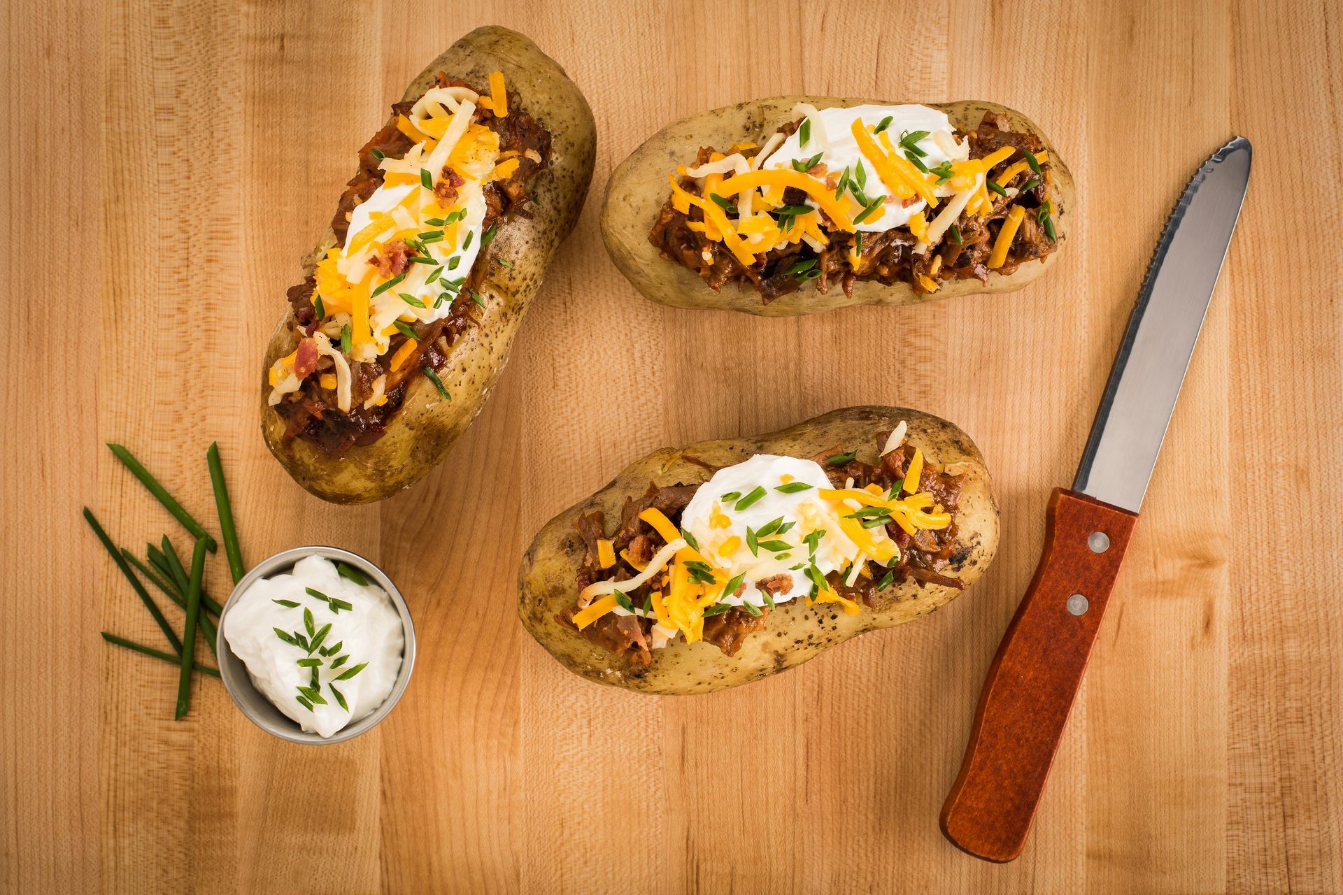 Baked potatoes topped with chili, cheese, sour cream, and chives on a wooden board.