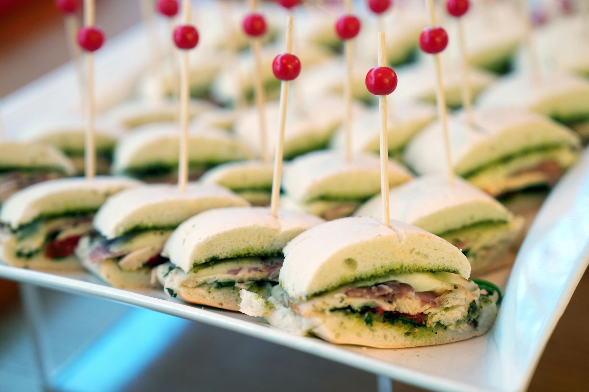 Mini sandwiches with pesto and red toothpick toppers on a white platter.