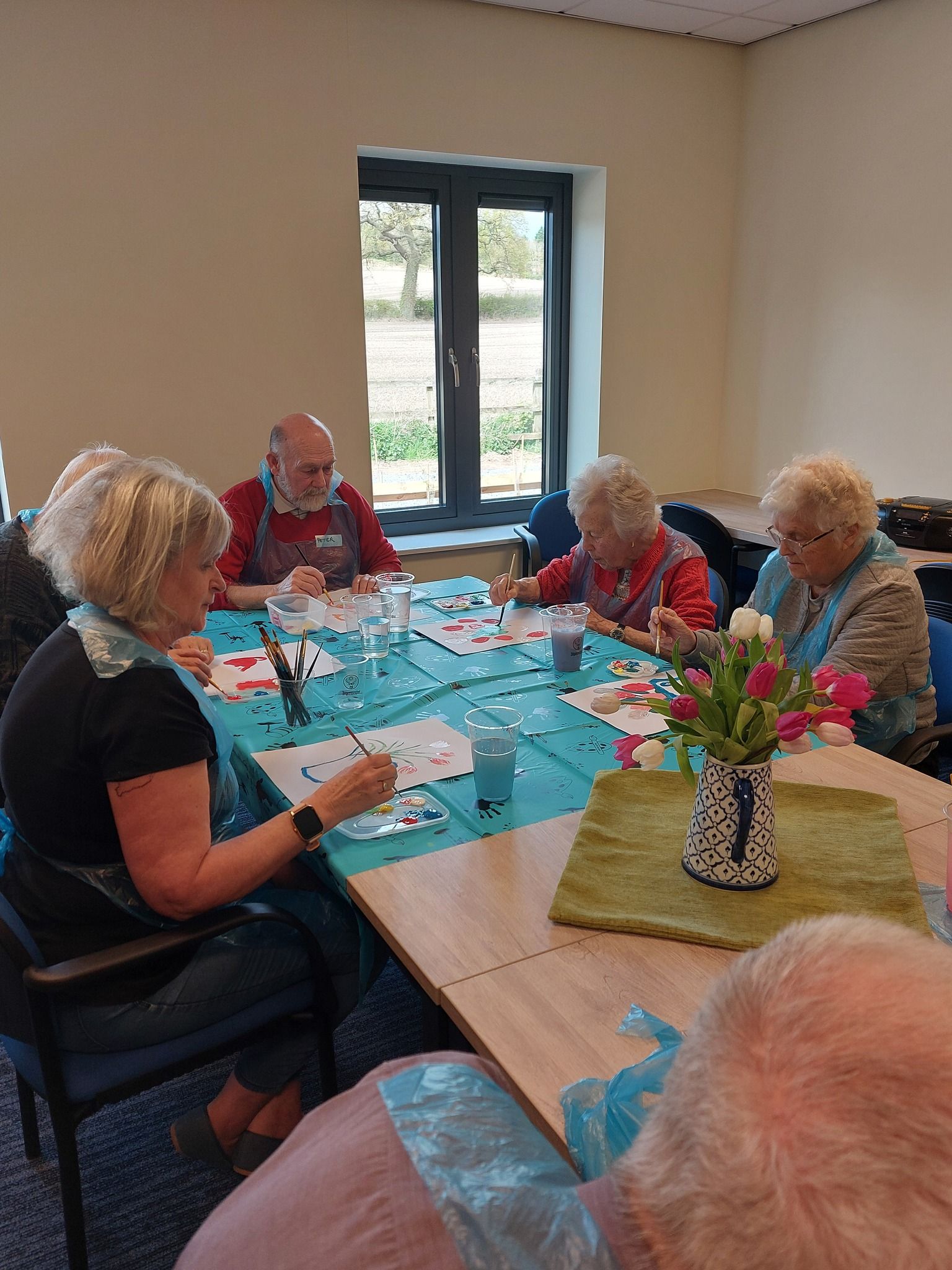 People seated around a table painting; blue tablecloth, flowers, window in the background.