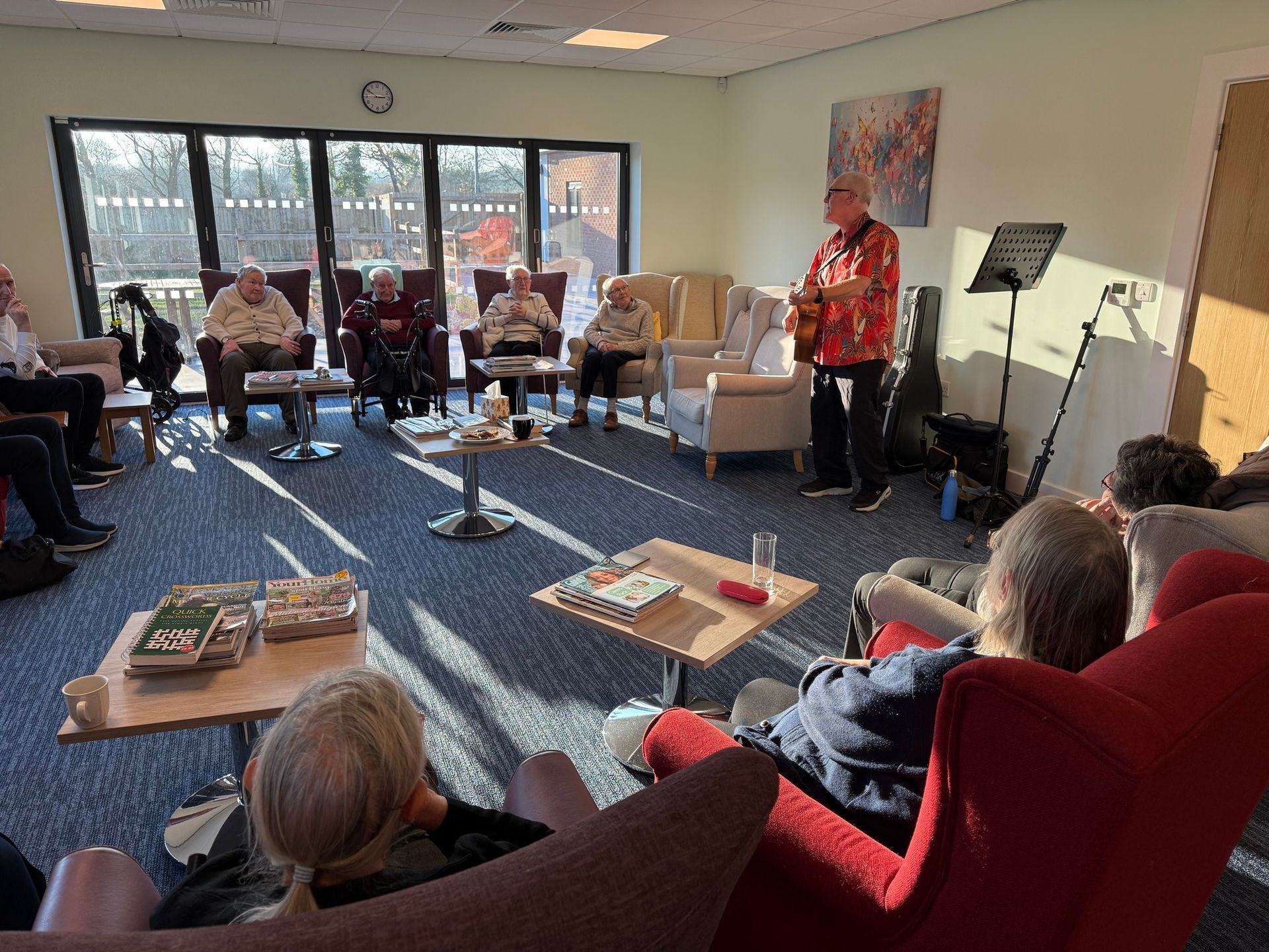 Group of people in armchairs listening to a person standing, possibly performing. Sunlight streams in from windows.