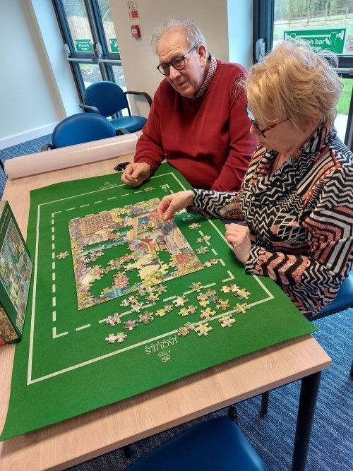 Two people working on a jigsaw puzzle at a table; green mat, indoor setting.