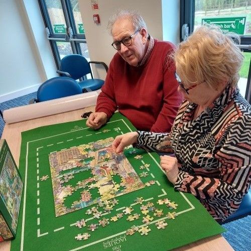 Two people seated, doing a jigsaw puzzle on a green mat. One points at a piece. Indoors, window, rolled paper.