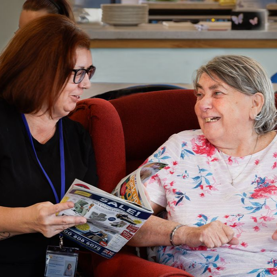 Woman smiles at an elderly person seated in a chair, a walker nearby.