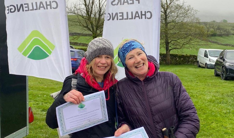 Two women smiling, holding certificates, posing in front of a banner at an outdoor event.