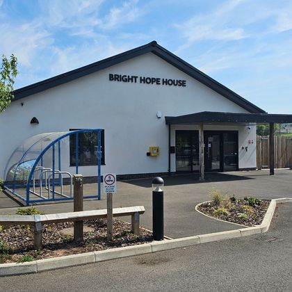 Four people sitting, watching something. One woman leans on a sofa, others are seated. White wall and sign in background.