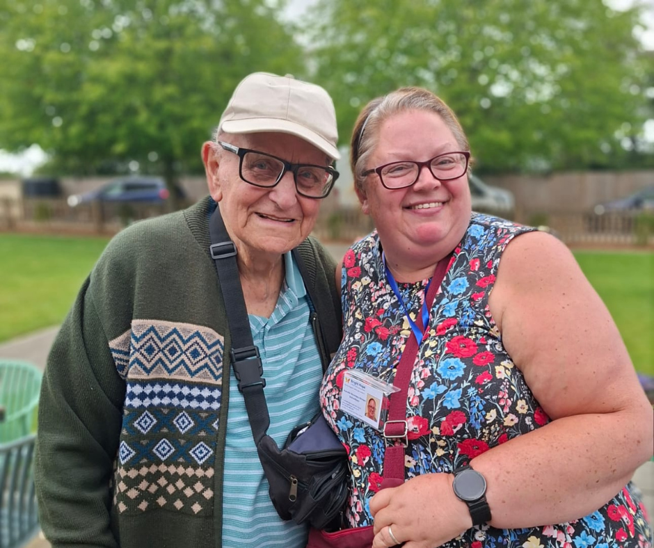 Woman talking to an older person in a care setting; both smiling, sitting. A walker is present.