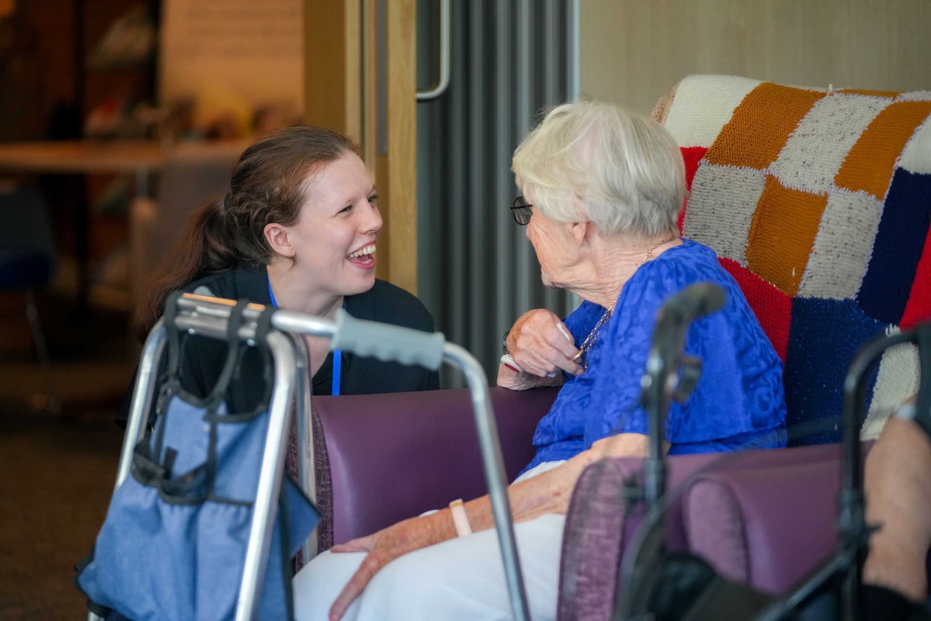 Woman talking to an older person in a care setting; both smiling, sitting. A walker is present.