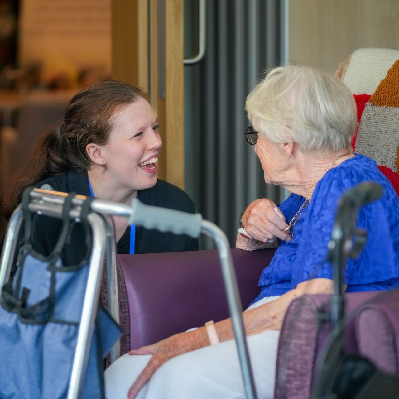 Woman smiles at an elderly person seated in a chair, a walker nearby.