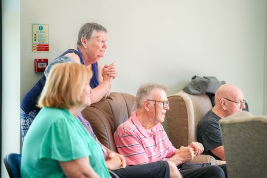 Four people sitting, watching something. One woman leans on a sofa, others are seated. White wall and sign in background.
