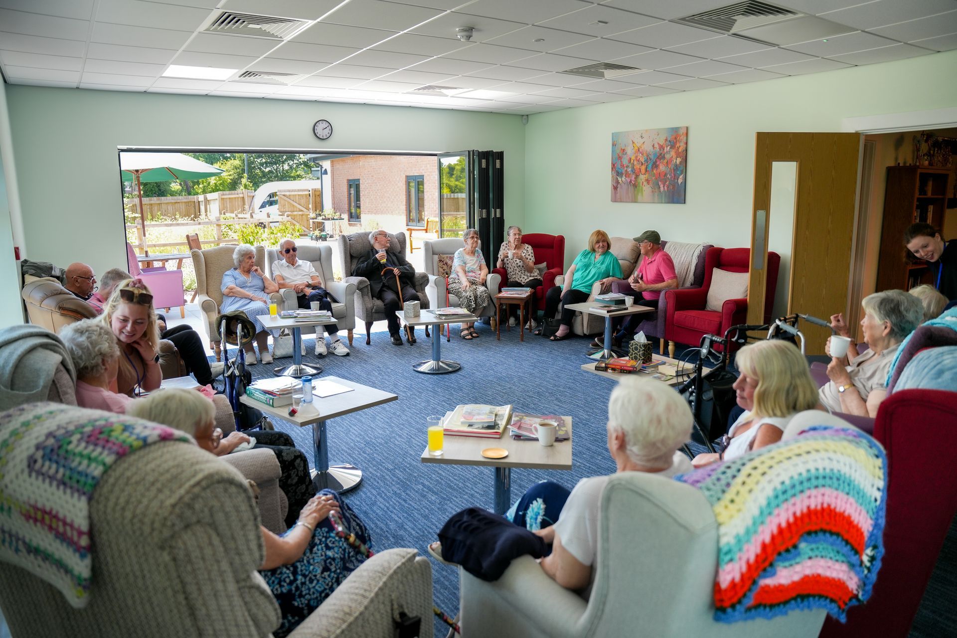 People seated in a circle, socializing in a bright room with open doors to a garden.