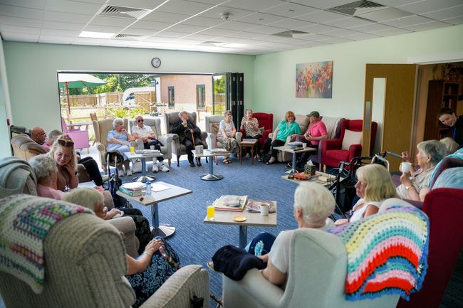 People seated in a circle, socializing in a bright room with open doors to a garden.