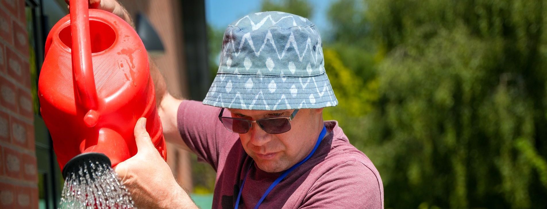 Person in a patterned hat and sunglasses watering plants with a red watering can outdoors.