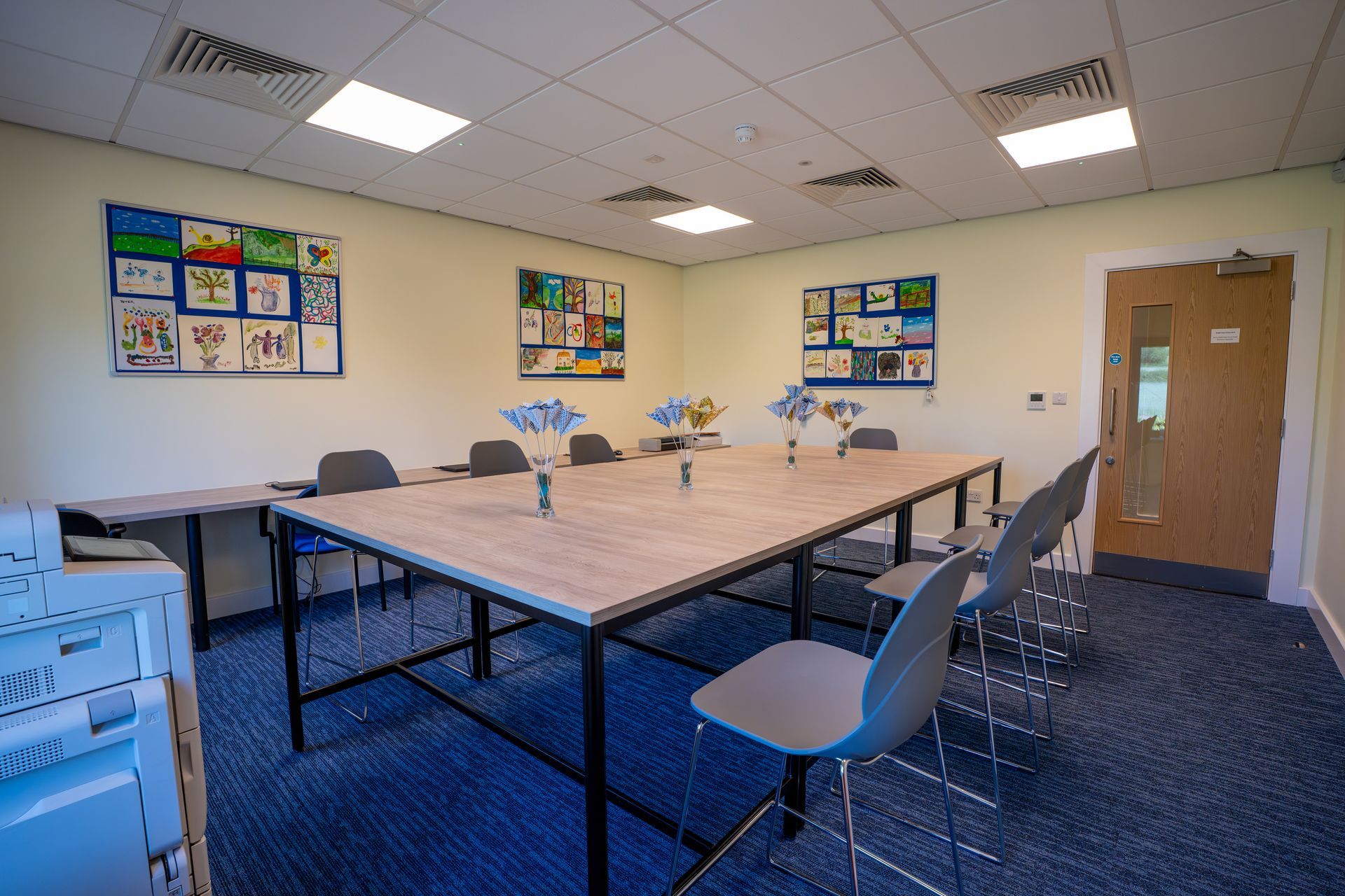 A brightly lit room with a large table, chairs, artwork on the walls, and a printer.