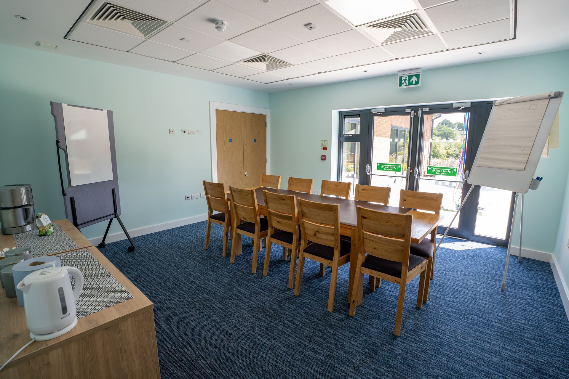 Meeting room: blue carpet, wooden table with chairs, two whiteboards, door to outside.