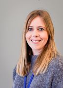 Woman with blonde hair smiles, wearing a grey sweater, in front of a grey background.