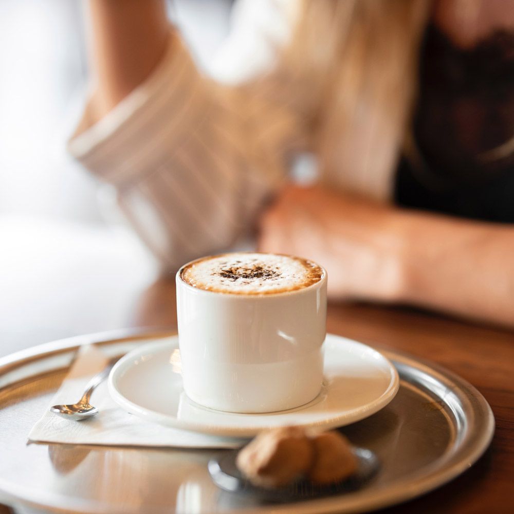 Cup of cappuccino on a silver tray, with a person in the background, near a window.