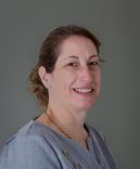 Woman with brown hair in a blue shirt smiling against a gray background.