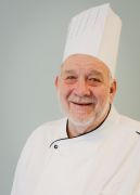 Chef smiling, wearing white chef's hat and coat, against a light blue background.