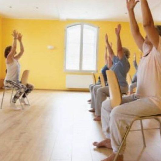 People in chairs doing exercises with arms raised in a sunny room.