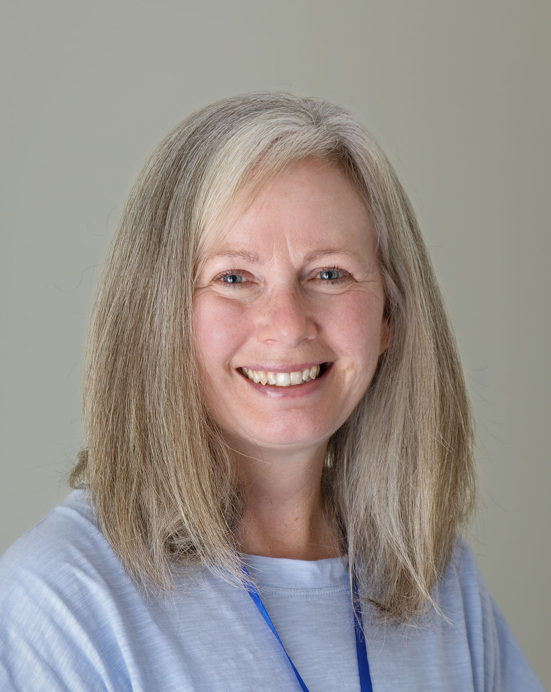 Woman with long, graying hair smiling, wearing a light blue shirt, against a pale background.