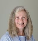Woman with long, graying hair smiling, wearing a light blue shirt, against a pale background.