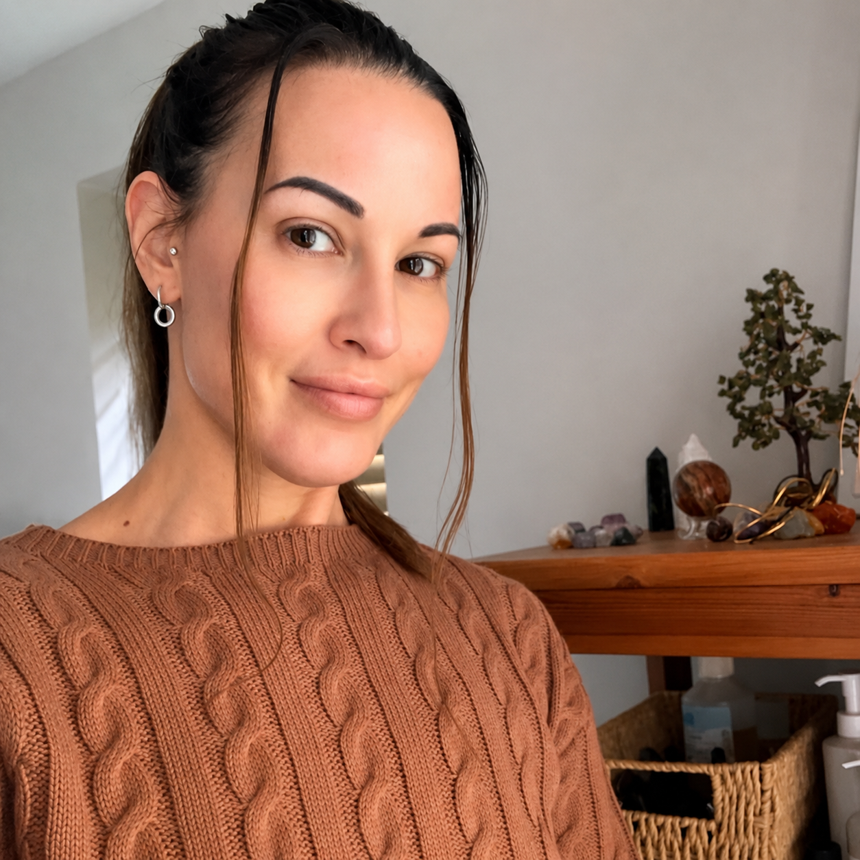 Woman in a brown sweater posing indoors beside a wooden table with plants and decor.