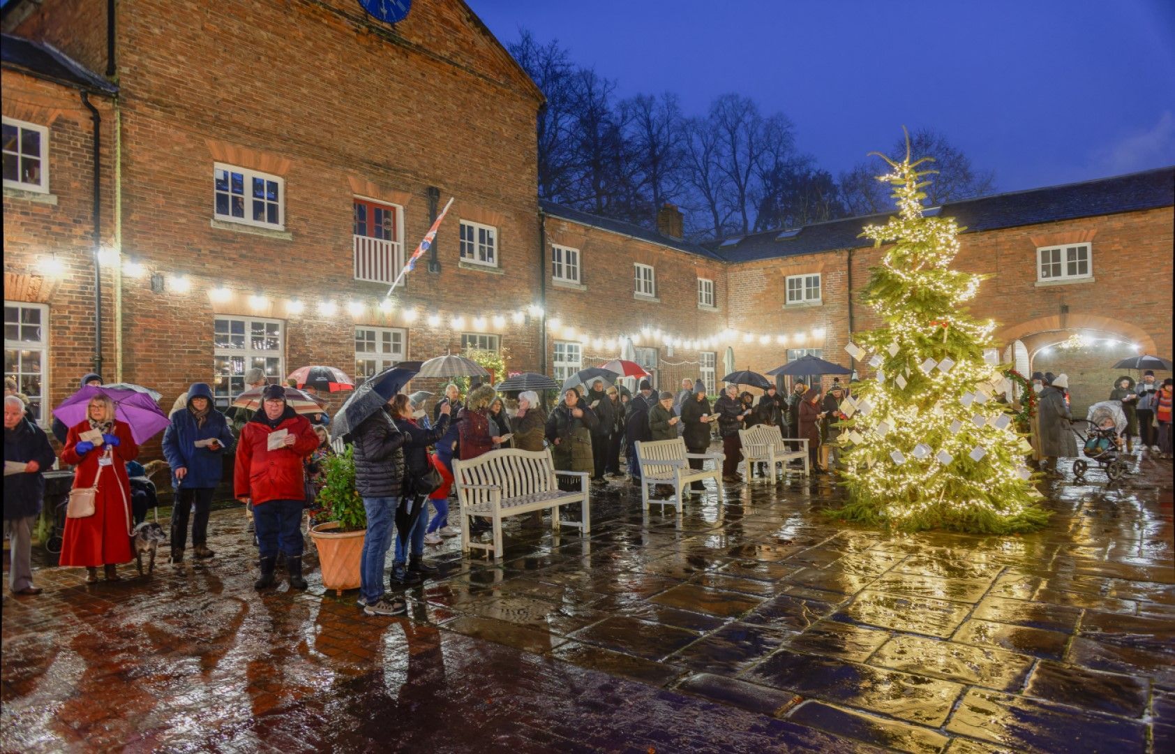Christmas tree and crowd gather outdoors, bricks buildings in the background. Rain reflects lights.
