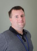 Man wearing a gray shirt with a black collar and a blue lanyard, smiling against a neutral background.