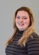 Woman with long blonde hair smiles, wearing a striped gray turtleneck top, against a gray background.