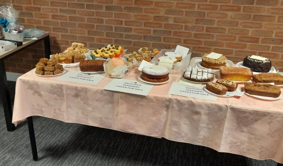 A table displays various baked goods like cakes and bars for sale, set against a brick wall.