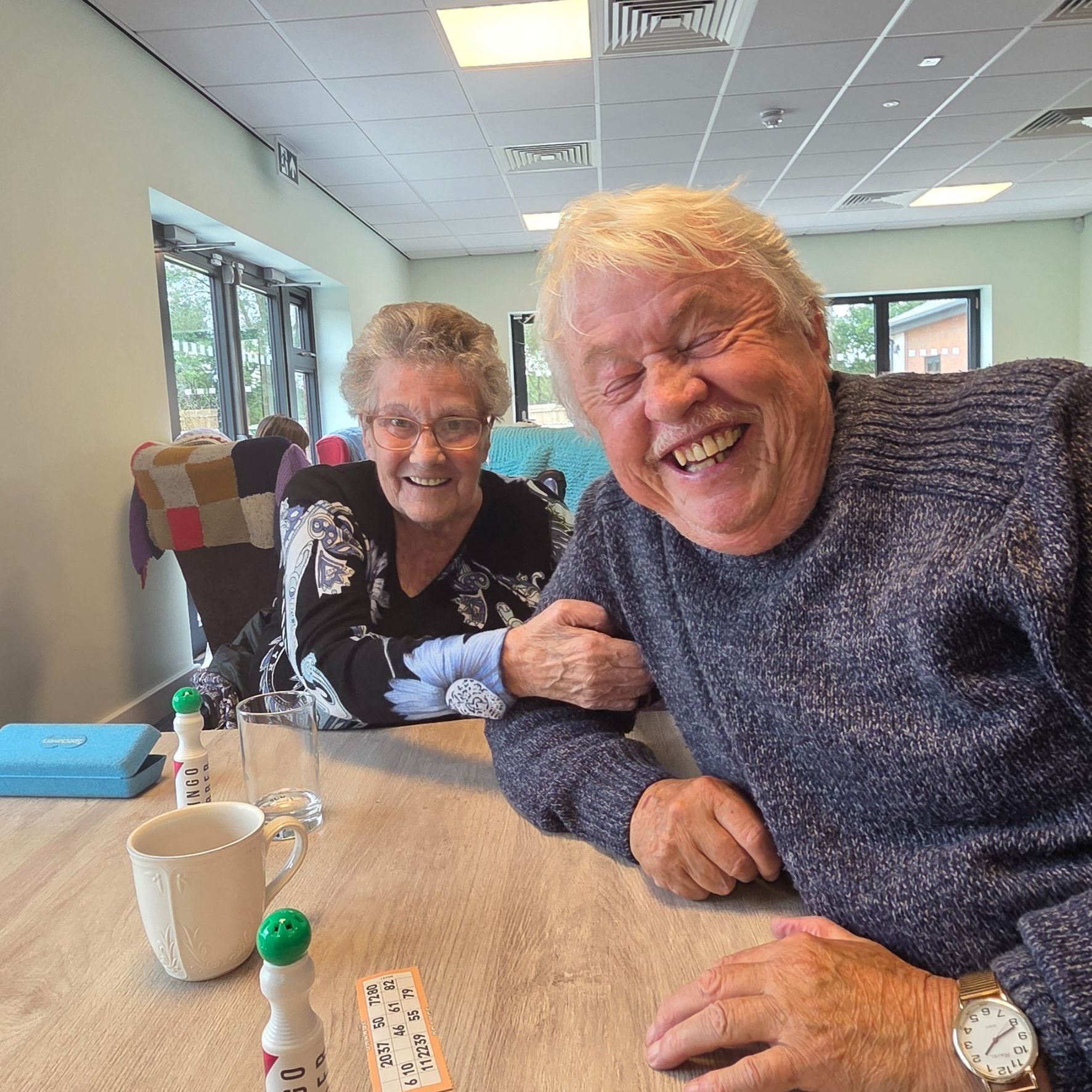 Two people working on a jigsaw puzzle at a table; green mat, indoor setting.