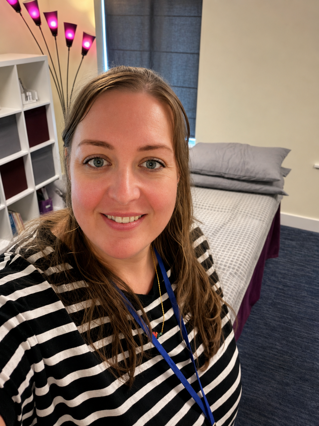 Person smiling in a striped sweater, taking a selfie in a bedroom with a bed and soft pink lamp lights.